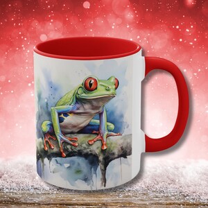 May include: A white ceramic mug with a red rim and handle. The mug features a watercolor painting of a green tree frog with red eyes sitting on a branch.