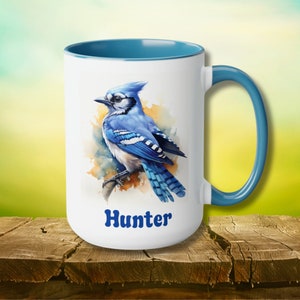 May include: White ceramic mug with a blue rim and handle. The mug features a watercolor illustration of a blue jay with a blue and white background. The text "Hunter" is printed in blue on the mug.