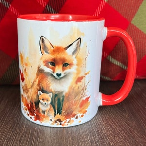 May include: White ceramic mug with a red interior and handle. The mug features a watercolor illustration of a mother fox and her cub surrounded by autumn leaves. The fox has orange fur and a white chest.
