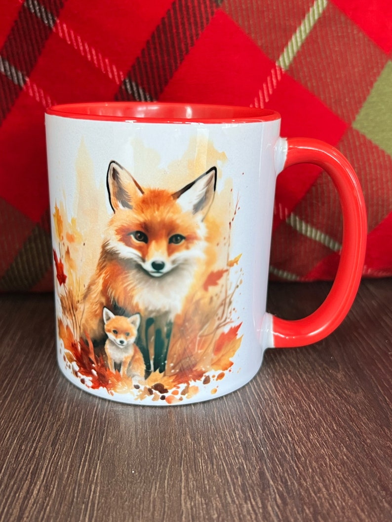 May include: White ceramic mug with a red interior and handle. The mug features a watercolor illustration of a mother fox and her cub surrounded by autumn leaves. The fox has orange fur and a white chest.