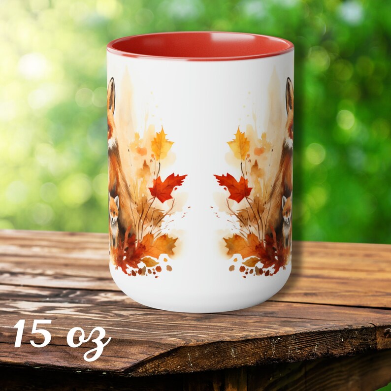 May include: A white 15 oz ceramic mug with a red interior. The mug features a watercolor design of a fox and autumn leaves in shades of orange, brown, and yellow. The mug is set on a wooden surface.