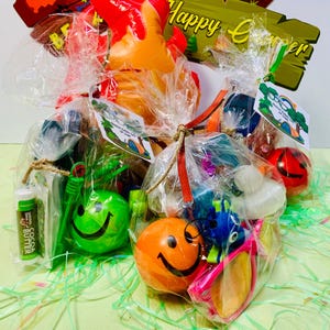May include: Gift baskets filled with various items, including smiley face balls, sunglasses, and lip balm. The baskets are wrapped in clear cellophane and tied with twine and ribbon. A wooden sign with the words "Happy Camper" is visible.