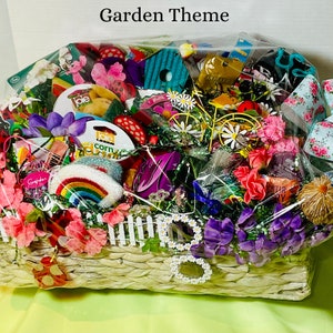 May include: A wicker basket filled with a variety of garden-themed items, including flowers, a rainbow, a miniature fence, and a small toy bicycle. The basket is wrapped in clear plastic and has a white label that says "Garden Theme".