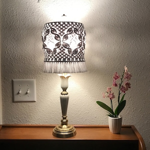 May include: A white lamp with a brown crocheted lampshade with a fringe. The lamp is on a wooden table with a white light switch on the wall behind it. There is a potted orchid with pink flowers on the table.