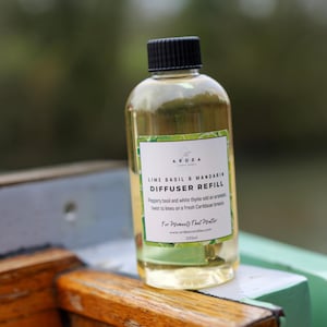 Könnte beinhalten: Eine klare Glasflasche mit Diffusor-Nachfüllung und schwarzem Deckel. Auf dem Etikett steht "Lime Basil & Mandarin Diffuser Refill" mit zusätzlichem Text. Die Flüssigkeit im Inneren ist gelb. Die Flasche steht auf einer Holzoberfläche.