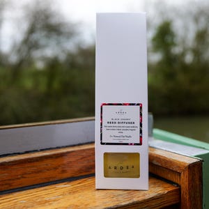 Può includere: Scatola rettangolare bianca per un diffusore a lamelle "Black Cherry" di Ardea, con un'etichetta floreale. La scatola è su una superficie di legno con un bordo verde, con uno sfondo sfocato di alberi e acqua.