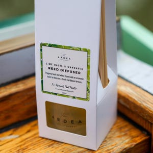 Op de afbeelding: Een witte doos met een rietverspreider en houten stokjes. Het etiket luidt "Lime Basil & Mandarin Reed Diffuser" met een groen en wit ontwerp. De doos staat op een houten oppervlak.