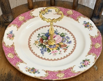 Op de afbeelding: Een serveerstandaard met één laag, bestaande uit een wit porseleinen bord met een centraal bloemenboeket, een roze en gouden decoratieve rand en een goudkleurig metalen handvat in het midden om het te dragen of uit te stallen.