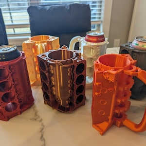 Peut inclure: Cinq mugs imprimés en 3D en forme de blocs moteur de différentes couleurs : rouge, orange, marron, or et blanc. Les mugs sont conçus pour contenir une canette de boisson.