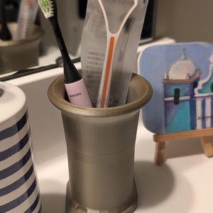 Puede incluir: Un soporte gris para cepillos de dientes con un cepillo de dientes Philips negro y rosa y un raspador de lengua en un paquete transparente. El cepillo de dientes tiene cerdas verdes. A la izquierda hay una taza de cerámica a rayas azules y blancas.