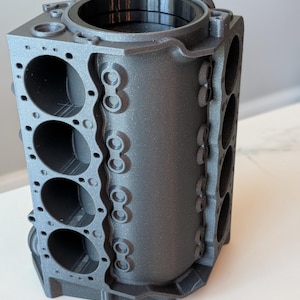 May include: A 3D printed metallic gray V8 engine block model featuring a detailed cylinder arrangement, bolt patterns, and a smooth black interior sleeve. The structure displays precise geometric cutouts and a textured finish throughout.