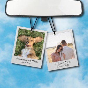 May include: Two hanging photo car air fresheners. The left one features a dog lying on its back with its tongue sticking out. The text below the photo reads "Personalized Photo And Text". The right one features a couple standing in a field. The text below the photo reads "I Love You, Drive Safe".