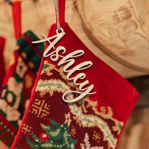 May include: A red Christmas stocking with a wooden name tag that says "Ashley" in cursive. The stocking has a detailed embroidered design with a Christmas tree and other festive patterns. A wooden mantel is in the background.