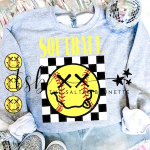 Può includere: Una felpa grigia con le parole "Softball" in lettere gialle e una palla da softball gialla con uno schema a scacchi nero e bianco dietro. La palla da softball ha un design cucito rosso e un viso sorridente con X per gli occhi.