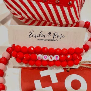 May include: Three red beaded bracelets with white letter beads spelling out the word "LOVE" in pink lettering. The bracelets are displayed on a white card with the text "Emilia Rose - HANDMADE JEWELRY -" printed on it.