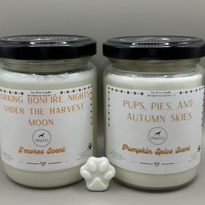 Puede incluir: Dos tarros de vidrio con velas de cera de soja con tapas negras y etiquetas blancas. Las etiquetas dicen "Barking Bonfire Nights Under The Harvest Moon" y "Pups, Pies, and Autumn Skies". Las velas tienen aroma a "S'mores" y "Pumpkin Spice". Hay un derretido de cera con forma de pata delante.