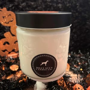 May include: A white candle in a decorative glass jar with a black lid. The jar has a label with a dog silhouette and the text "MILEY SUE'S CANDLE CO." The candle sits on a white pedestal, with Halloween decorations in the background.