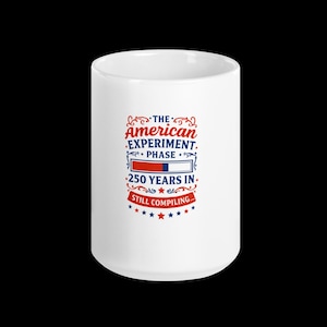 May include: White ceramic mug with the text "The American Experiment. Phase. 250 Years In. Still Compiling..." in red, white, and blue. The mug has a glossy finish and a simple, cylindrical shape.