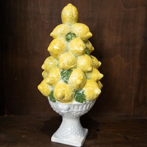 May include: A decorative ceramic topiary sculpture shaped like a pyramid of yellow lemons with green leaves, resting in a white pedestal bowl with a textured basket-weave pattern on the base and stem, standing upright on a flat square base.