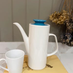 May include: A white ceramic coffee pot with a teal lid and a matching white mug. The coffee pot has a long spout and a handle. The mug is cylindrical with a small handle. The items are on a yellow book.