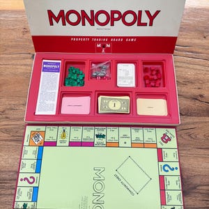 May include: A vintage Monopoly board game set with a red and white box. The box is open, revealing game pieces, cards, and play money. The game board is visible below, featuring property spaces and instructions.