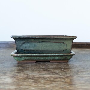 May include: A rectangular ceramic bonsai pot with a matching drip tray, featuring a mottled green and brown glaze. The pot has a wide, flared rim and a recessed panel on the side, showcasing a rustic, earthy texture with visible clay edges.