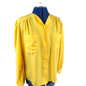 May include: A bright yellow button-down blouse with long sleeves and two front pockets. The blouse is displayed on a blue dress form against a white background. The fabric appears to be a lightweight material.