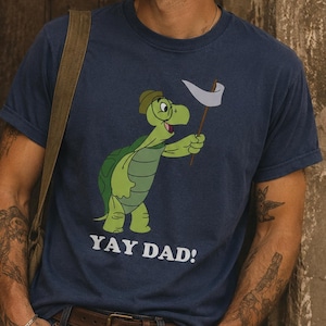 May include: A navy blue short-sleeve t-shirt featuring a graphic of a green turtle wearing a hat and holding a white flag. Below the turtle, the text YAY DAD! is printed in white capital letters. The shirt has a crew neckline and a casual fit.