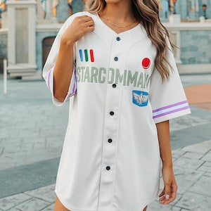 Op de afbeelding: Wit honkbalshirt met zwarte knopen, paarse strepen op de mouwen en de tekst "STARCOMMAY". Voorzien van een rode cirkel en een Buzz Lightyear-patch. Het shirt wordt gedragen voor een kasteel.