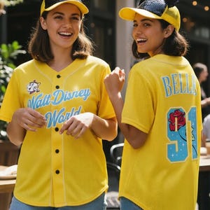 May include: Two people wearing yellow baseball jerseys with "Walt Disney World" in light blue script. They are also wearing yellow caps with black sequined mouse ears. The jersey has black buttons and a cartoon graphic.