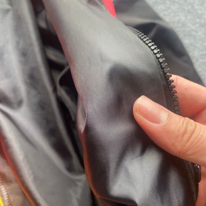 May include: Close-up of the black lining inside a red and black jacket. The lining is smooth and has a subtle sheen. A zipper is visible along the edge of the lining.