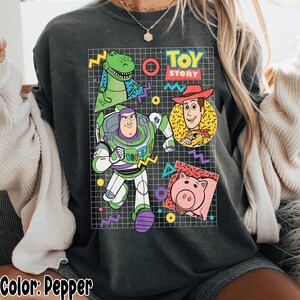 May include: Pepper-colored sweatshirt featuring a graphic of Toy Story characters: Rex, Buzz Lightyear, Woody, and Hamm. The design includes the Toy Story logo and geometric shapes on a grid background. The text "Color: Pepper" is at the bottom.
