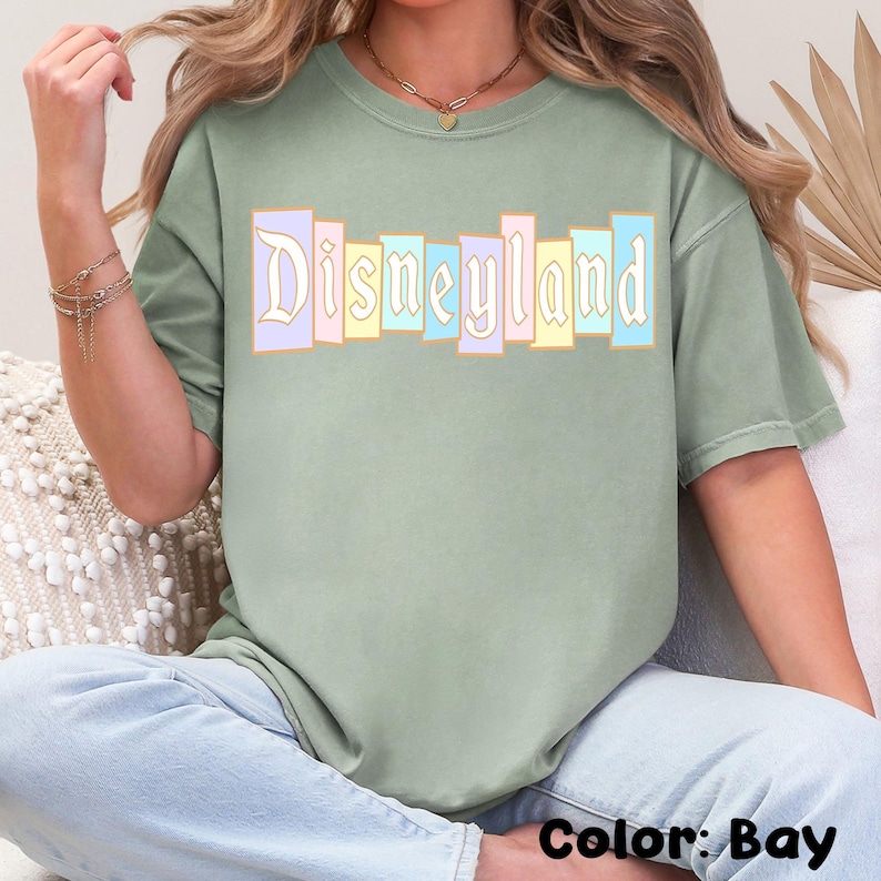 Puede incluir: Camiseta verde salvia con la palabra "Disneyland" en un dise&ntilde;o de bloques de color pastel. El texto est&aacute; en una fuente vintage. La camiseta es de corte holgado y el color es "Bay".