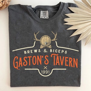 Op de afbeelding: Een donkergrijs T-shirt met een vintage-stijl logo met een bruine ton met een kraan, geweien en de tekst "Brews & Biceps Gaston's Tavern 1991".