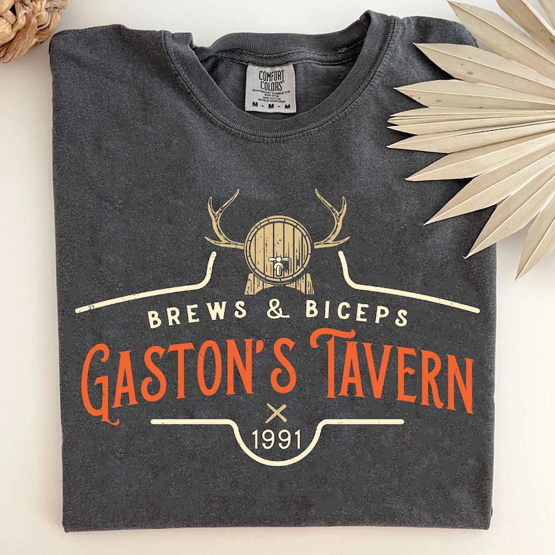 Op de afbeelding: Een donkergrijs T-shirt met een vintage-stijl logo met een bruine ton met een kraan, geweien en de tekst "Brews & Biceps Gaston's Tavern 1991".