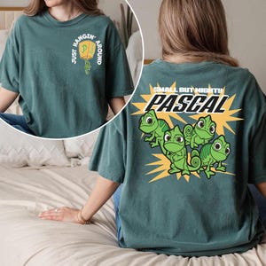 Puede incluir: Camiseta de color verde azulado con el texto "Just Hangin' Around Pascal" en la parte delantera y "Small But Mighty Pascal" con gráficos de camaleones verdes en la parte trasera. La camiseta tiene un corte holgado.