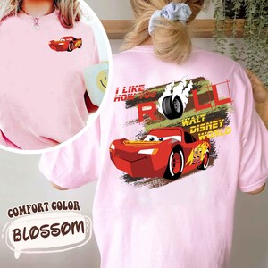 Puede incluir: Camiseta rosa claro con un dise&ntilde;o de coche de carreras rojo de dibujos animados en la parte delantera y trasera. El dise&ntilde;o trasero incluye el texto "I LIKE HOW YOU ROLL" y "WALT DISNEY WORLD". La camiseta est&aacute; etiquetada como "COMFORT COLOR BLOSSOM".