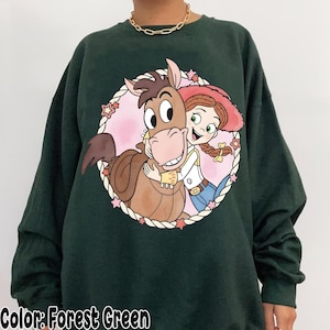 May include: Forest green sweatshirt featuring a cartoon graphic of a horse and a cowgirl hugging inside a pink circle with stars. The text "Color: Forest Green" is at the bottom of the image.