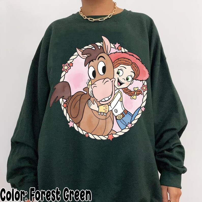 May include: Forest green sweatshirt featuring a cartoon graphic of a horse and a cowgirl hugging inside a pink circle with stars. The text "Color: Forest Green" is at the bottom of the image.