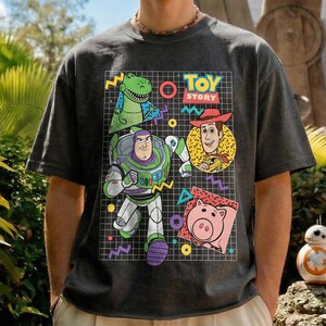 May include: Dark gray t-shirt featuring a colorful graphic of Toy Story characters. The design includes Buzz Lightyear, Woody, Rex, and Hamm, set against a grid background with geometric shapes. The words "TOY STORY" are also displayed.