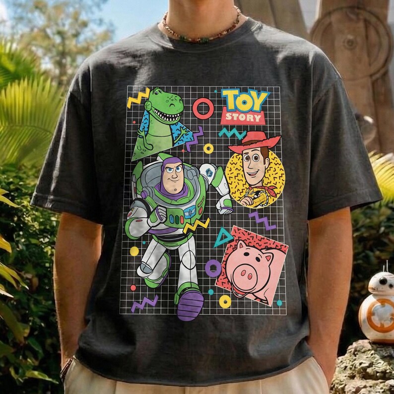 May include: Dark gray t-shirt featuring a colorful graphic of Toy Story characters. The design includes Buzz Lightyear, Woody, Rex, and Hamm, set against a grid background with geometric shapes. The words "TOY STORY" are also displayed.