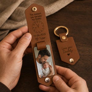 May include: Brown leather keychain and key fob set. The keychain has a photo of a father and baby, with the text "The moment you became my Daddy" and a date. The key fob says "Love you Daddy".