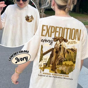 May include: Ivory-colored t-shirt with "EXPEDITION EVEREST" graphic on the back, featuring a mountain scene and the text "BE WARE OF THE YET!". The front has a small circular graphic. The shirt is from Animal Kingdom Park.