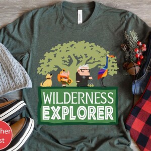 May include: A green t-shirt featuring a cartoon image of a character from the film Up. The character is standing next to a tree and a bird. The text "WILDERNESS EXPLORER" is printed on the shirt.