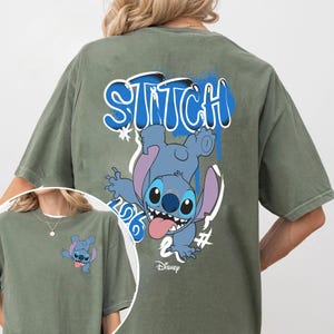 Puede incluir: Camiseta verde oliva con un personaje de dibujos animados azul y la palabra "Stitch" en letras azules estilo grafiti. El personaje tiene una gran sonrisa y saca la lengua. El logotipo de Disney est&aacute; en la parte inferior.