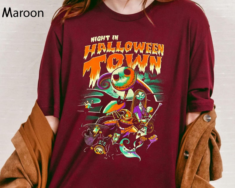 Może przedstawiać: Bordowy t-shirt z nadrukiem graficznym postaci z filmu "Koszmar przed świętami". Tekst na koszulce brzmi "Night in Halloween Town".