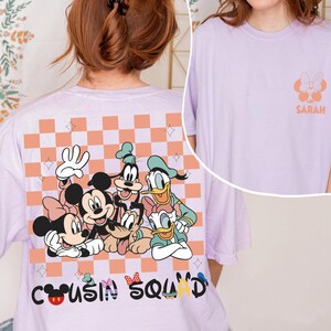 May include: A lilac t-shirt with a graphic of Disney characters, including Mickey and Minnie Mouse, Goofy, Donald and Daisy Duck. The back of the shirt has a checkered pattern and the words "Cousin Squad". The front has a small graphic and the name "Sarah".