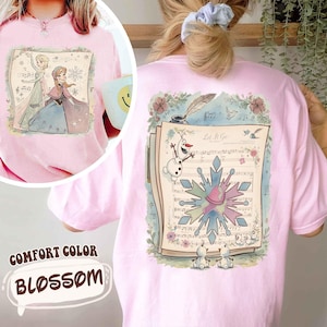 May include: Pink Comfort Color Blossom t-shirt featuring a watercolor illustration of Elsa and Anna from Frozen on the front. The back displays a musical score with Olaf and a snowflake design, with the words "Let It Go".