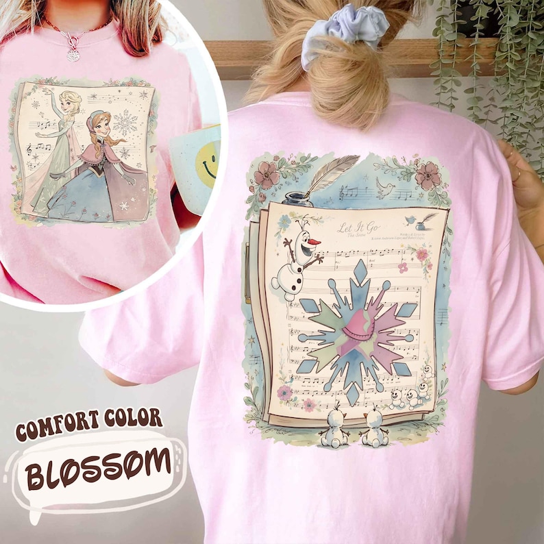 May include: Pink Comfort Color Blossom t-shirt featuring a watercolor illustration of Elsa and Anna from Frozen on the front. The back displays a musical score with Olaf and a snowflake design, with the words "Let It Go".