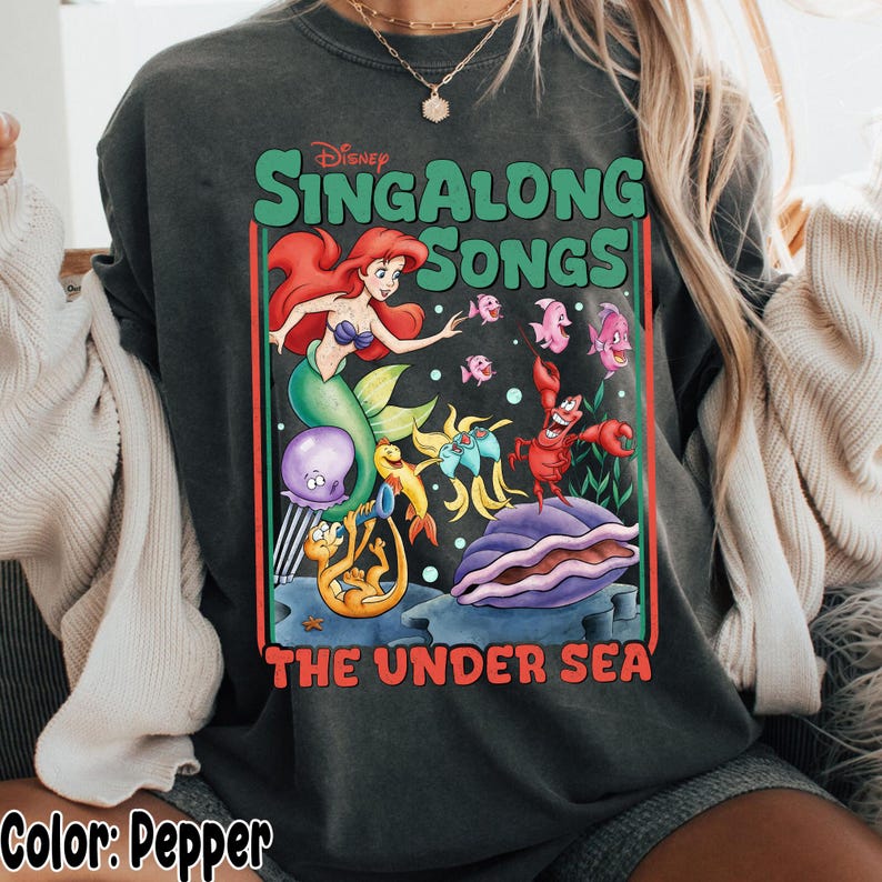 Pu&ograve; includere: Felpa color pepe con una grafica colorata di "Singalong Songs: The Under Sea" della Disney. Il design include Ariel, creature marine e il testo "Singalong Songs" e "The Under Sea".